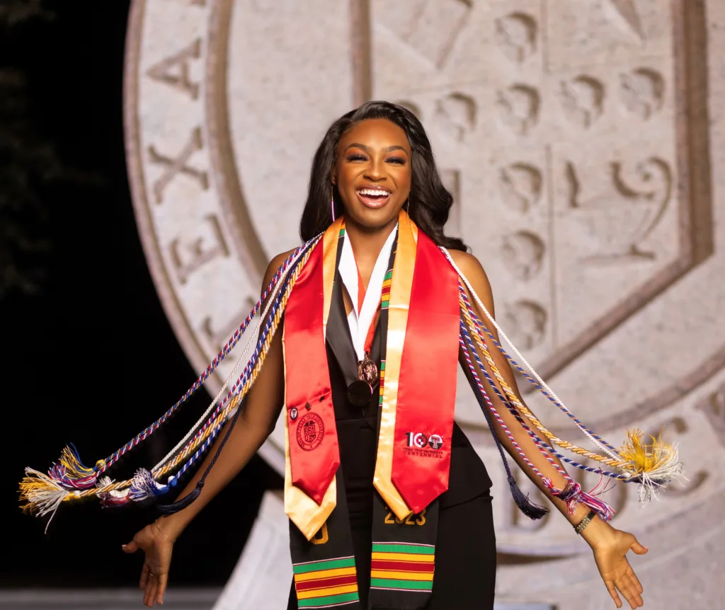 Christianah, A Joyful Graduate, Adorned With Multiple Colorful Honor Cords And A Kente Stole, Celebrates In Front Of A Large, Ornate Seal. - Best Charter School Texas Education