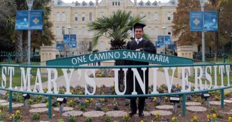 Giovanny Sanchez Graduating In Cap And Gown Smiling Proudly In Front Of A Semicircular Green Fence With &Quot;St Mary'S University&Quot; Sign, Backed By A Historic Building And Garden. - Best Charter School Texas Education