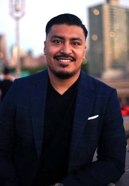 Giovanny Sanchez With A Warm Smile, Sporting A Neatly Trimmed Beard And Wearing A Dark Blue Suit With A White Pocket Square, Poses Outdoors During Dusk. - Best Charter School Texas Education