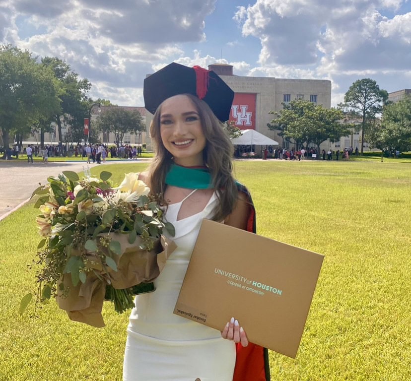 Dr. Tania Escobar Standing In The Sun, Wearing A White Dress And A Graduation Cap While Holding Flowers And A University Of Houston College Degree